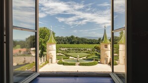 Uitzicht door een raam op een formele tuin met symmetrische beplanting, twee torens en een heldere lucht.