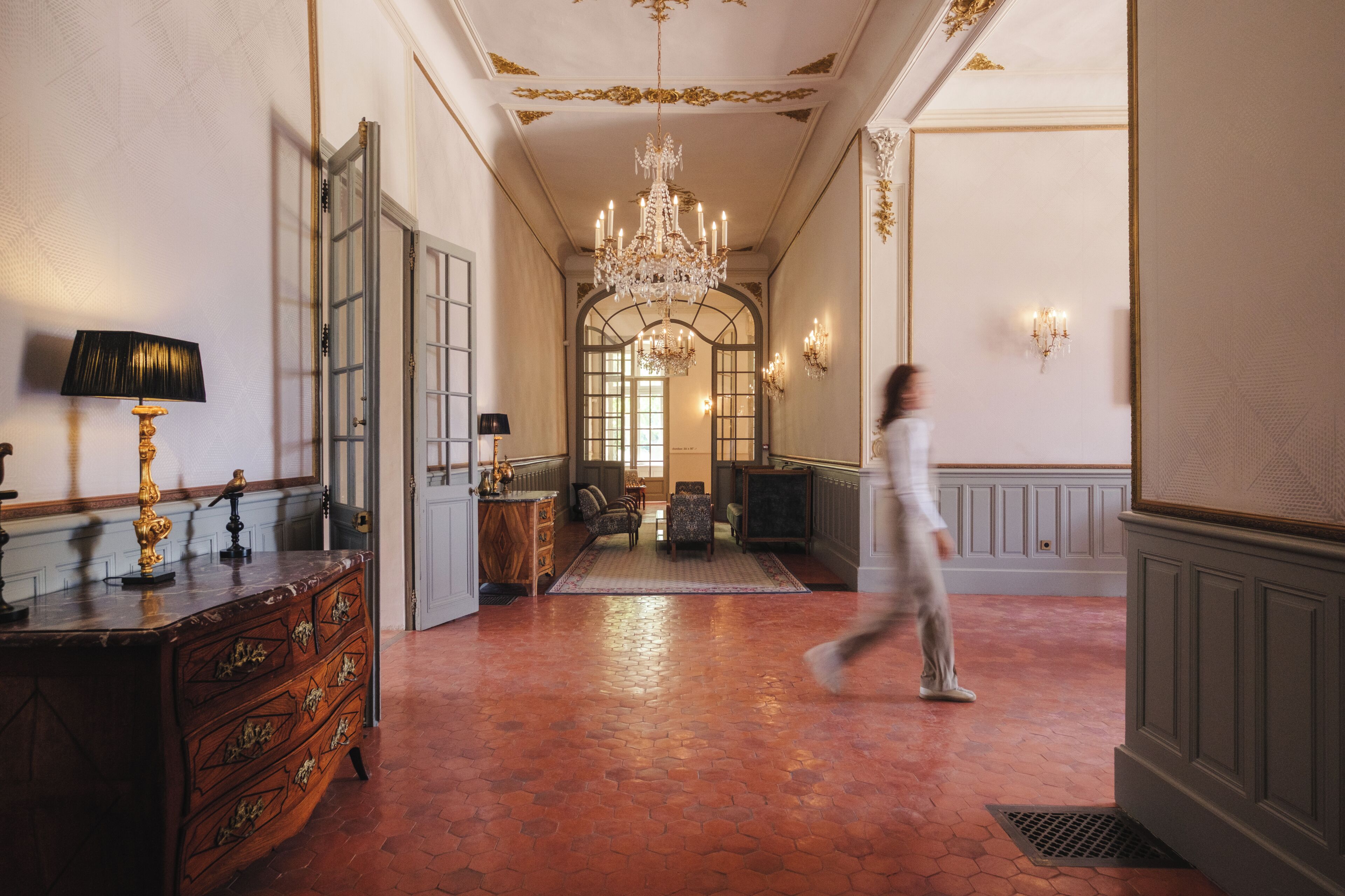 A hallway with a large chandelier, a wooden dresser, and a person walking.