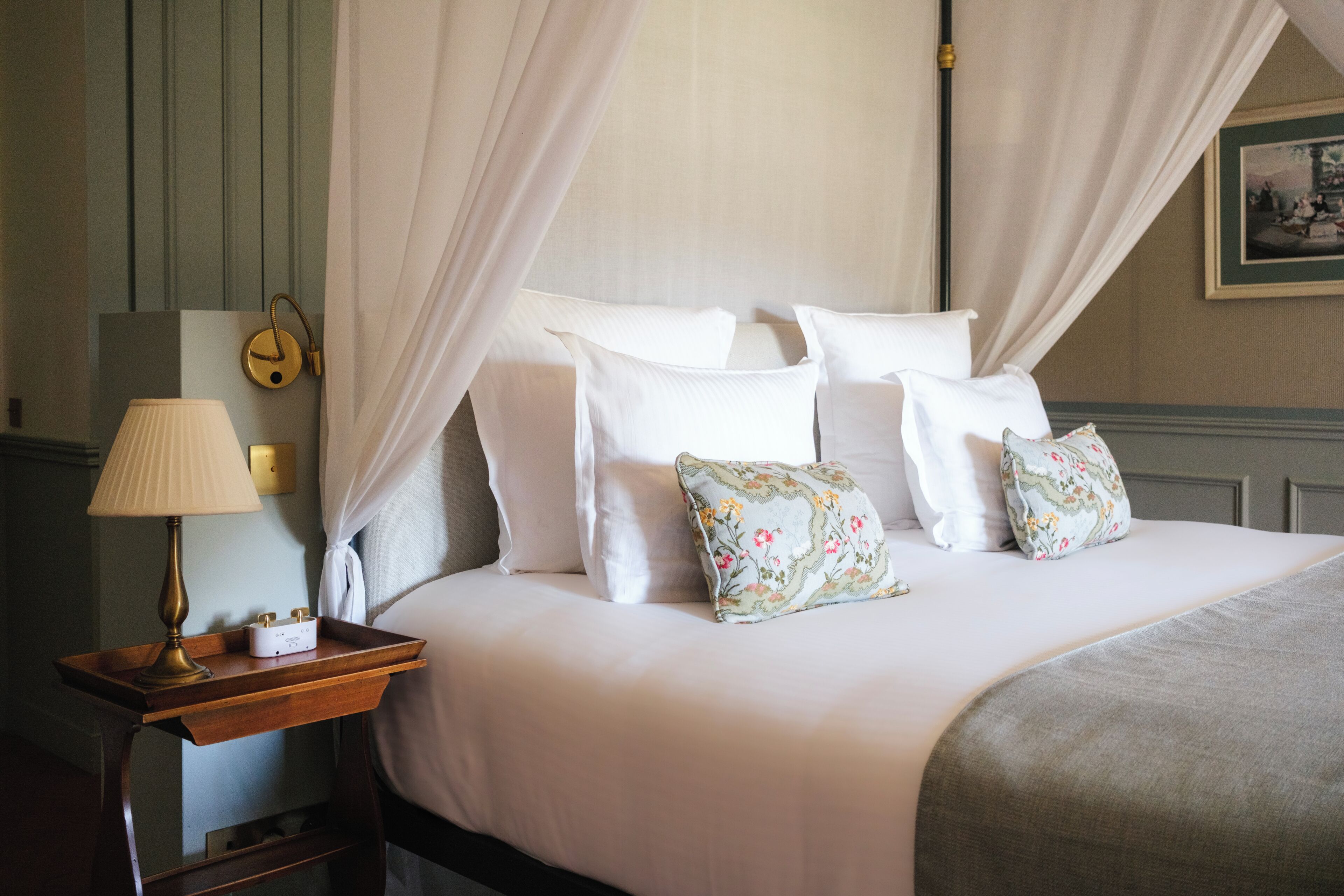 A neatly made bed with white linens and floral pillows, a bedside table with a lamp, and a framed picture on the wall.