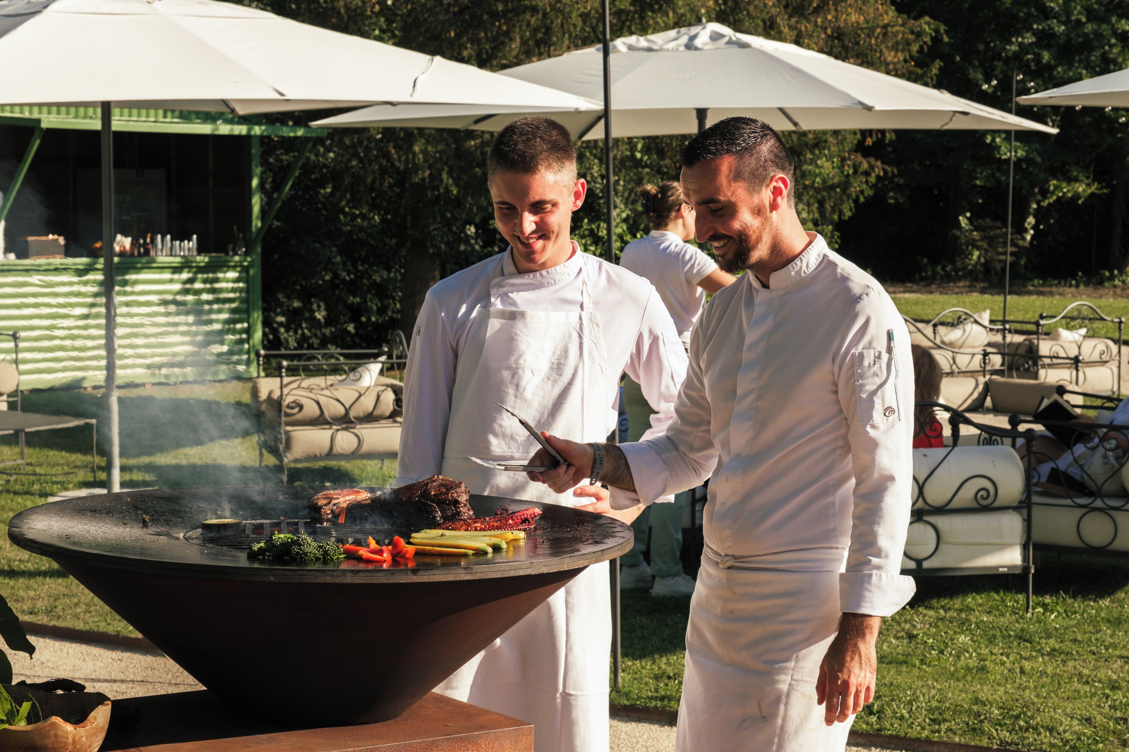 Zwei Köche bereiten Essen im Freien mit einem Grill zu, umgeben von Sonnenschirmen und viel Grün.