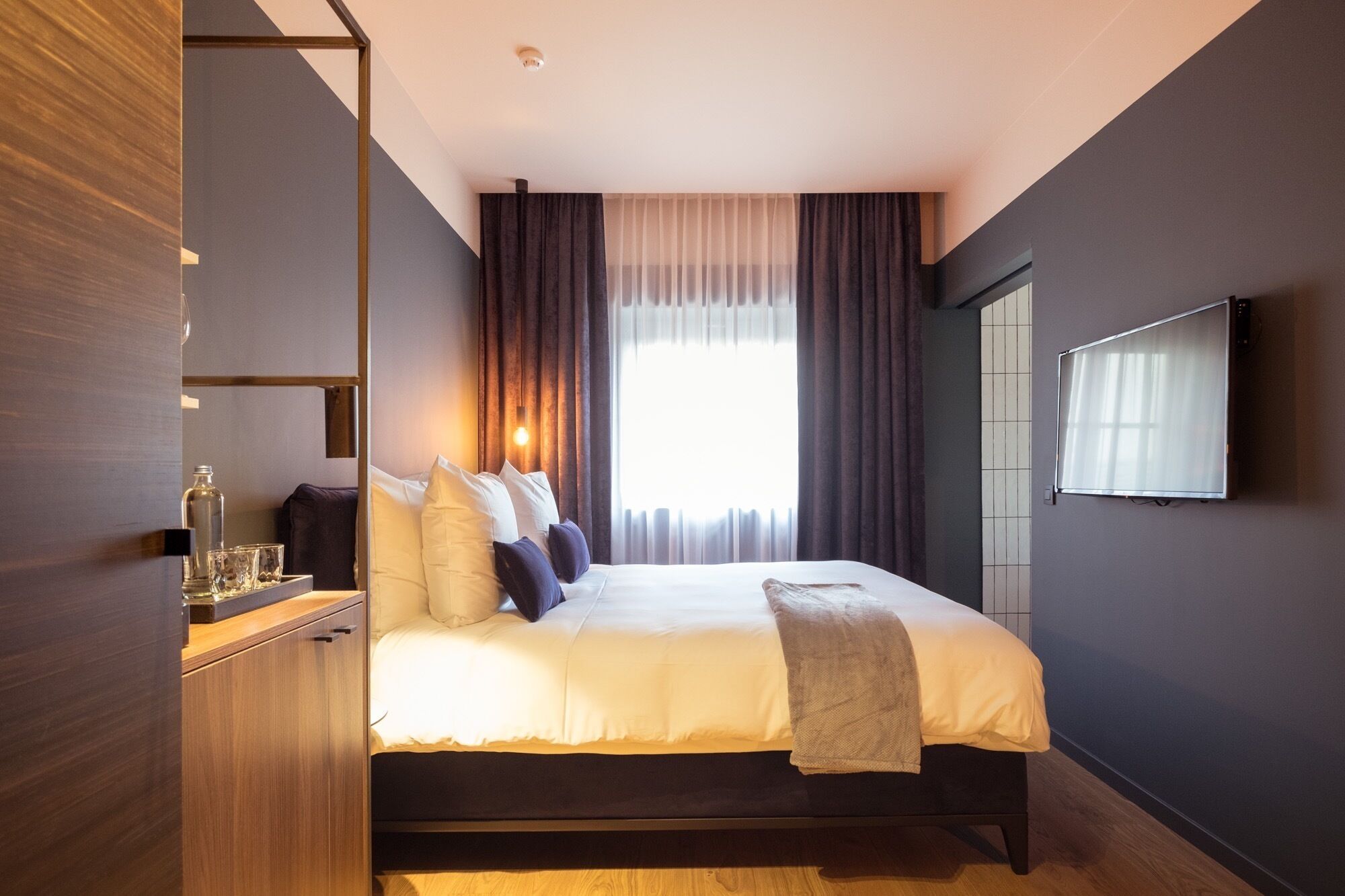 A modern hotel room with a large bed, a bedside table with glasses, a bottle, and a remote control.