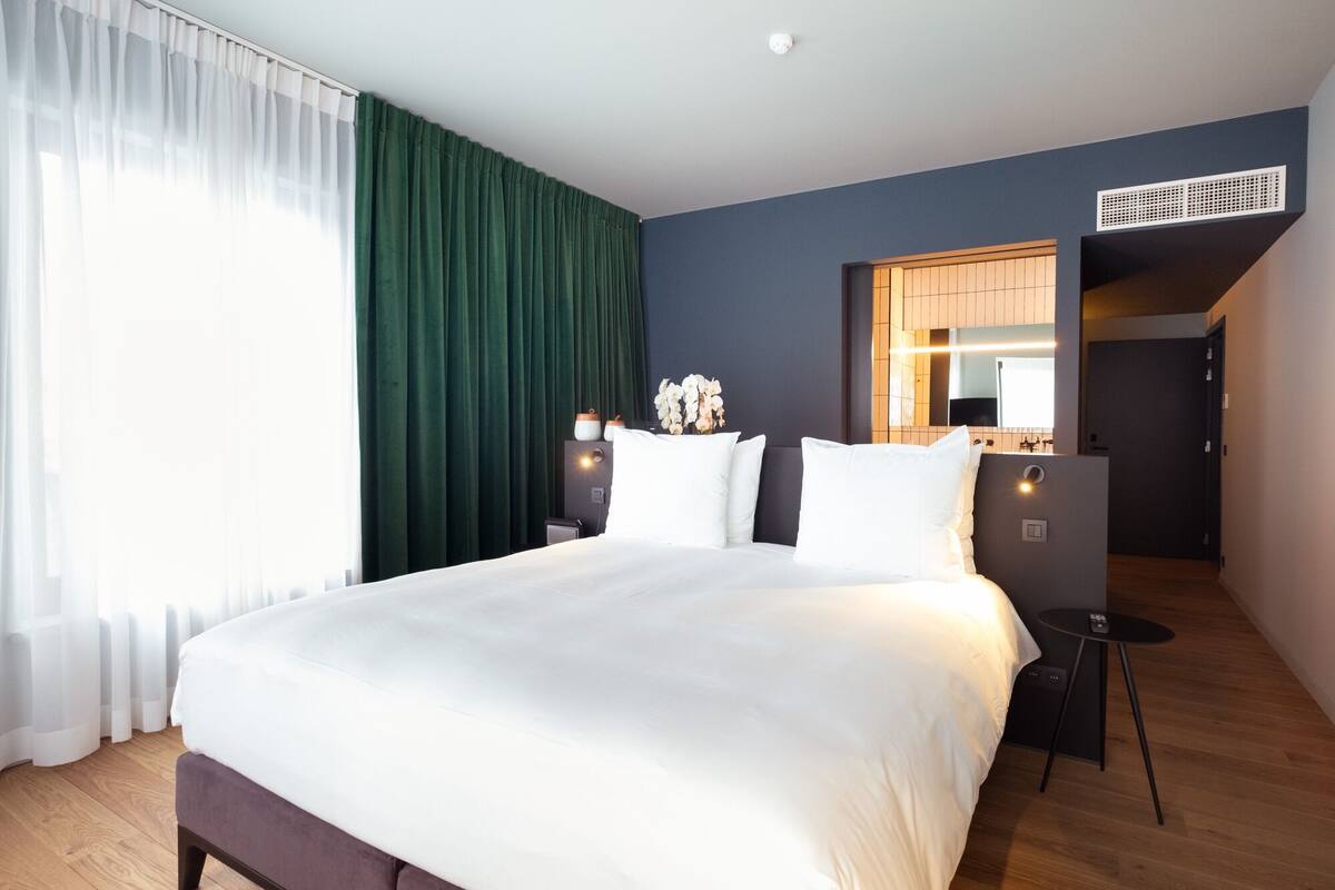 A modern hotel room with a large bed, green curtains, a small table, and a mirror.