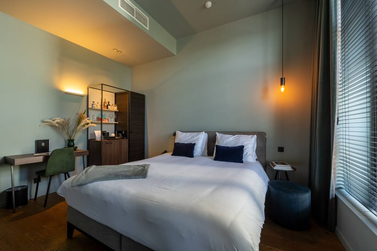 A modern hotel room with a large bed, a desk with a chair, a small table, and a window with blinds.