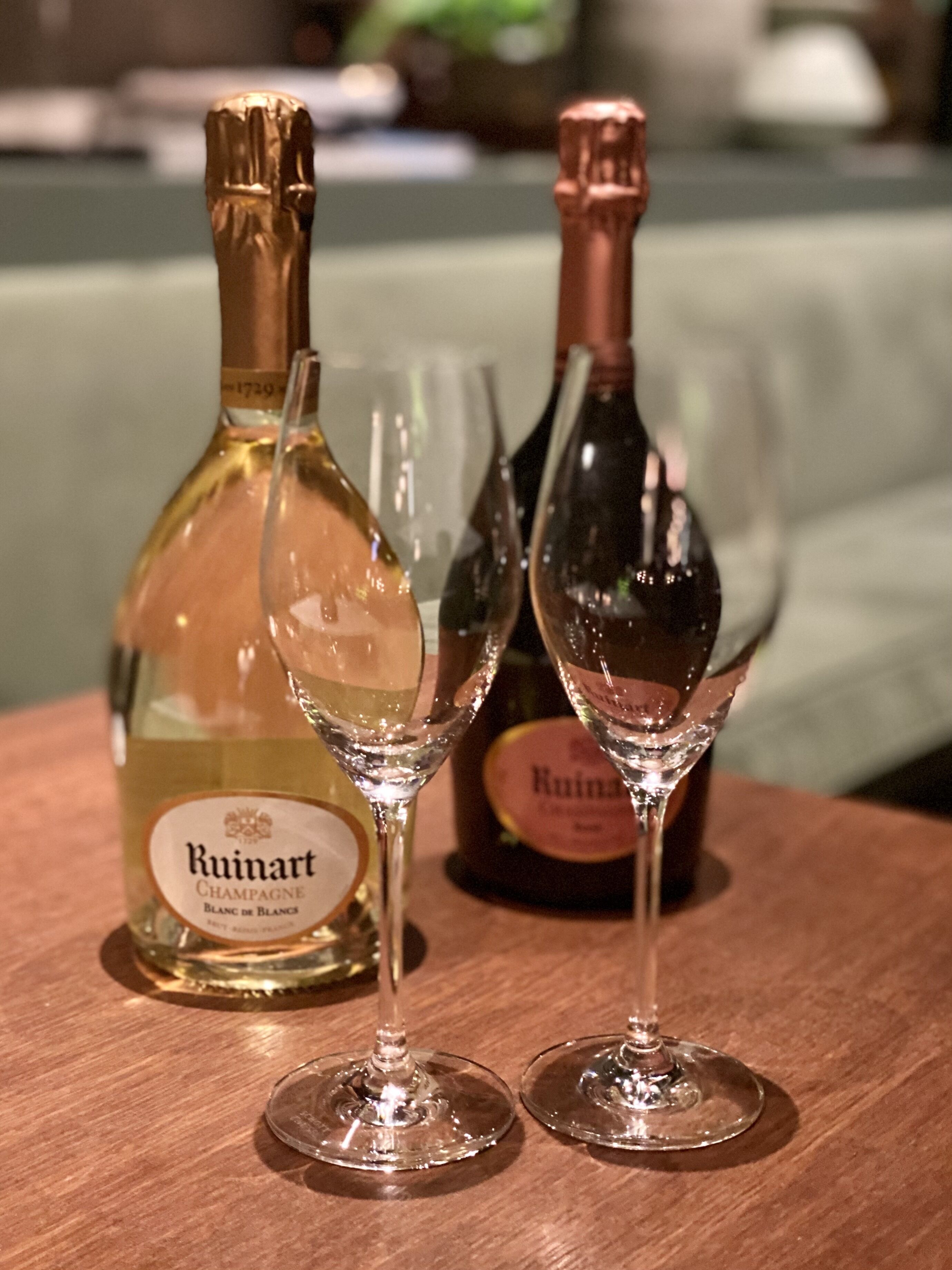 A bottle of Ruinart Champagne Blanc de Blancs and two glasses on a wooden table.