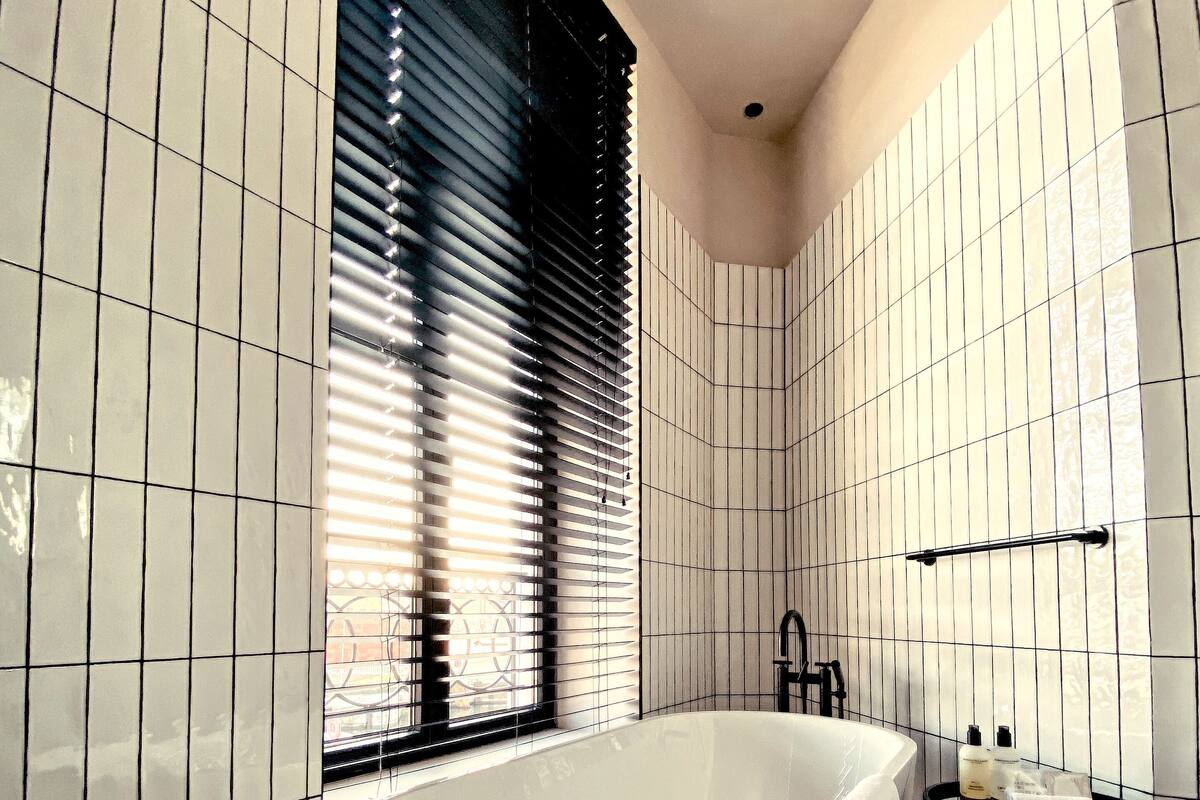 A modern bathroom with white tiled walls, a freestanding bathtub, a window with blinds, and a small black side table with toiletries.