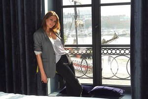 Una persona de pie junto a una ventana con vista a un puerto deportivo y barcos.