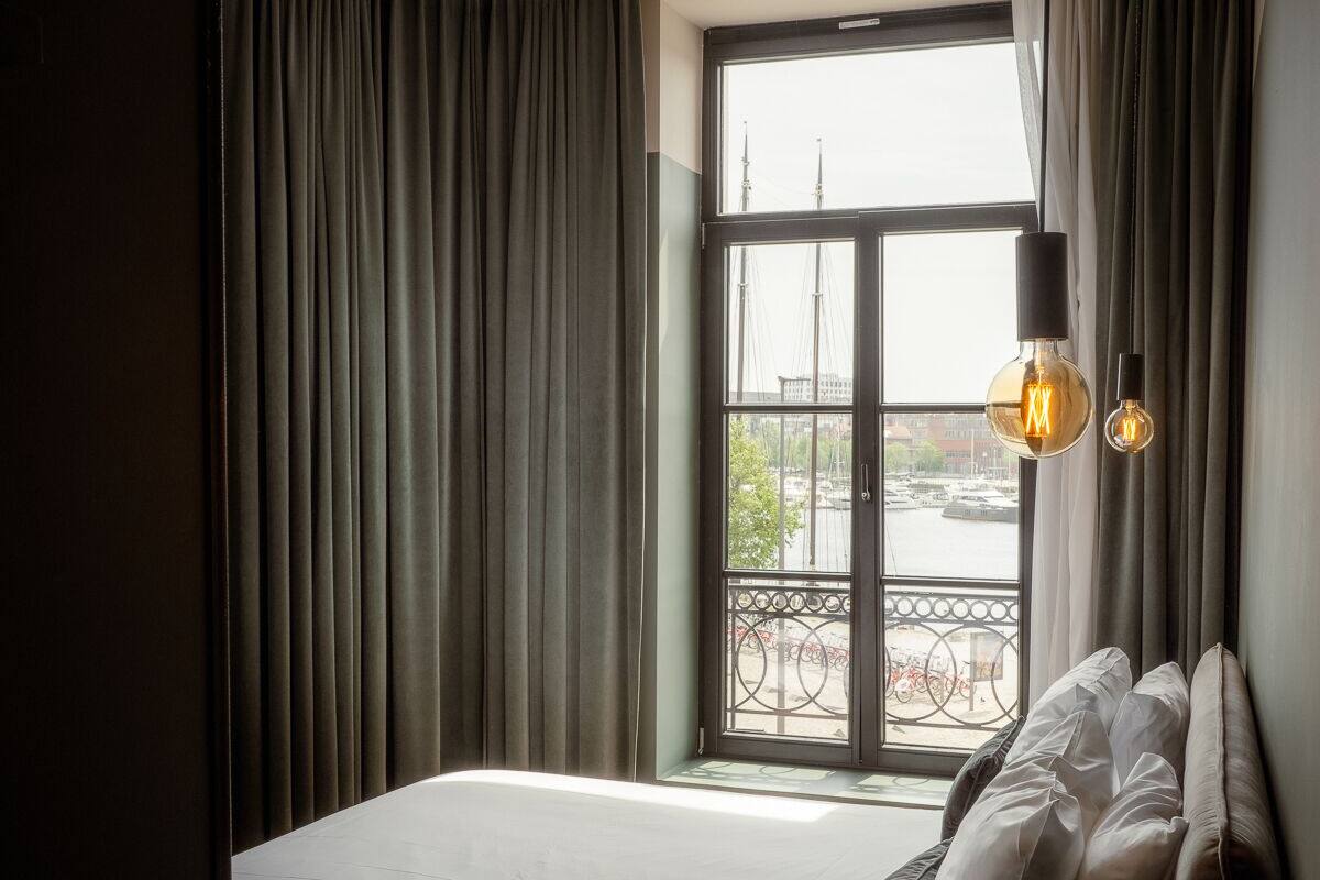 A modern bedroom with a large bed, a view of a cityscape through a window, and a balcony with a railing.