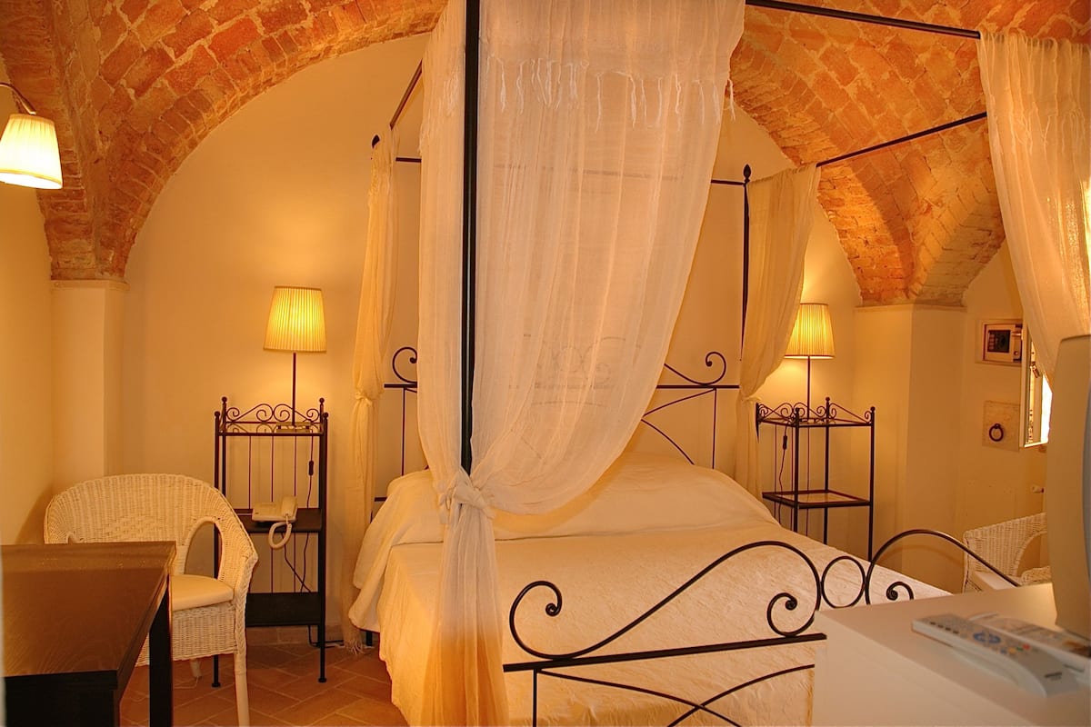 A four-poster bed with a canopy, a wicker chair, a bedside table with a lamp, and a telephone.