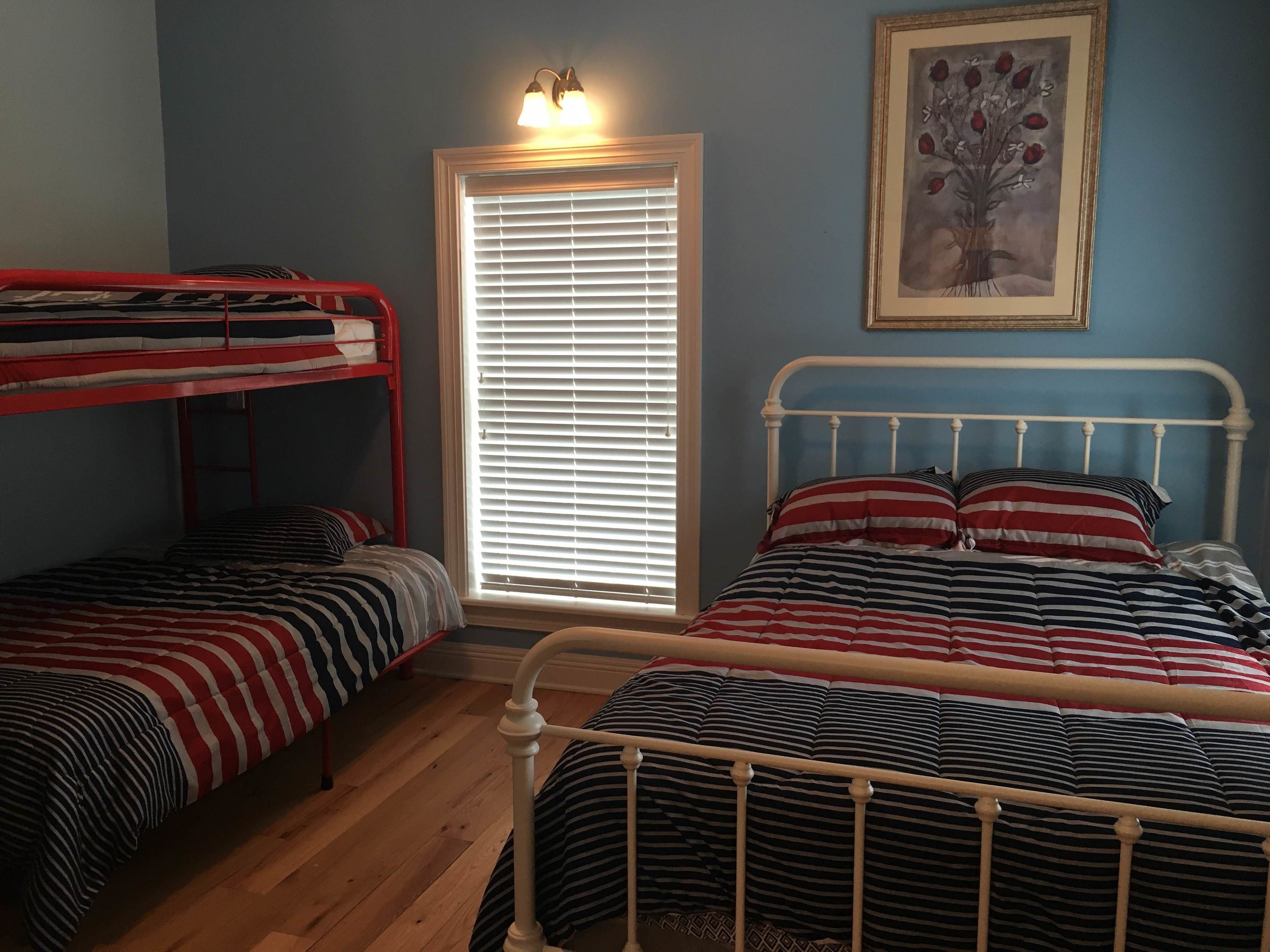 A room with two bunk beds, one with a red frame and the other with a white frame. There is a window with blinds, a framed picture on the wall, and a wooden floor.