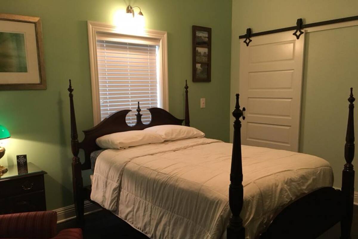 A bedroom with a four-poster bed, a nightstand with a lamp, a window with blinds, and a door.