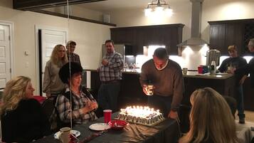 A group of people gathered around a table with a birthday cake, some holding drinks, in a room with dark wooden cabinets and a modern kitchen in the background.