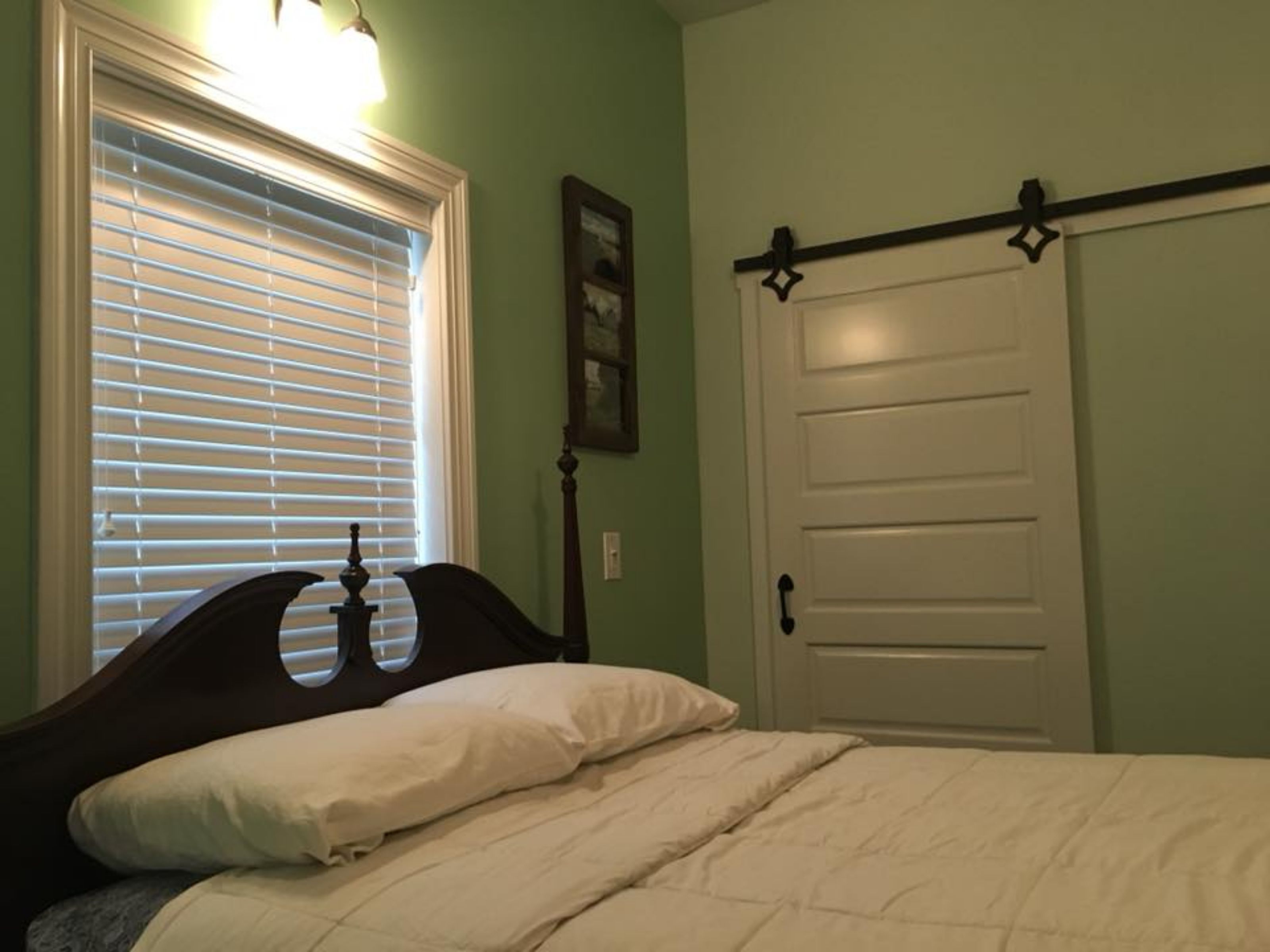 A bedroom with a bed, a window with blinds, a door with a black handle, and a wall-mounted picture.