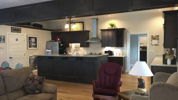 A spacious living area with a large sofa, a red armchair, and a coffee table. A kitchen with dark cabinets and a central island is visible in the background. - Crystal Run Suites of Springboro (Springboro)