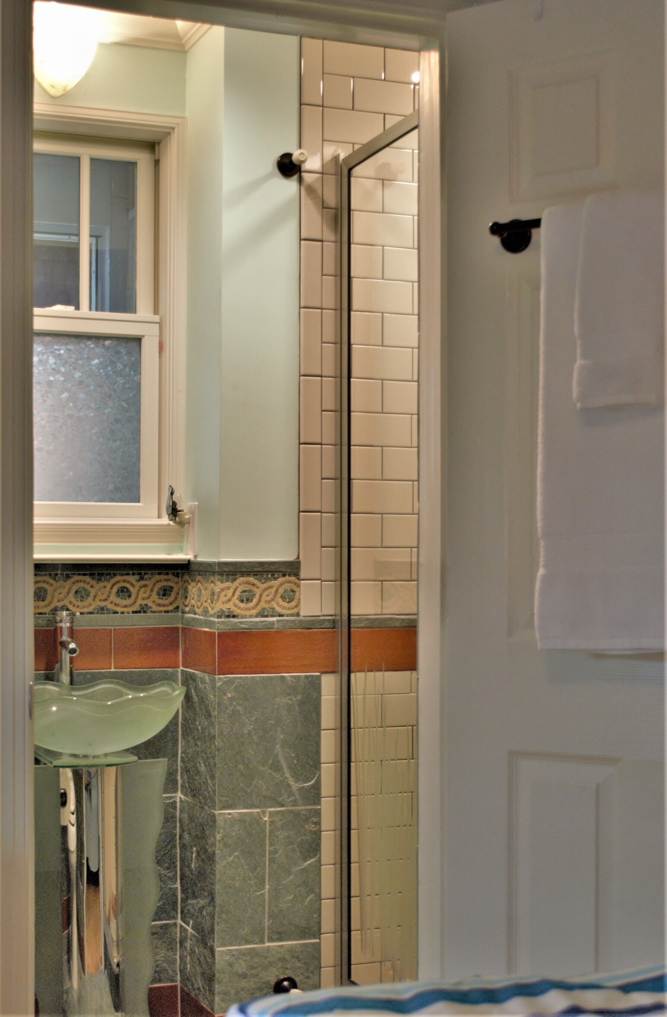 A bathroom with a glass shower enclosure, a sink with a greenish glass basin, and a tiled wall with a decorative border.