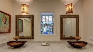 A bathroom with two sinks, each with a bowl sink and a mirror above. There is a window with a stained glass design between the mirrors. - The Grey Owl Inn (St. Simons Island)