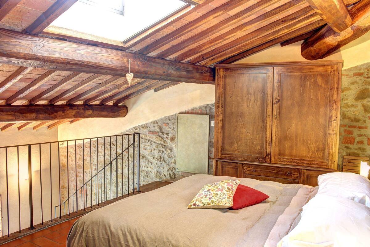 Ein Schlafzimmer mit schrägverlaufender Decke, Holzbalken, einem Bett mit einem Blumenkissen, einem hölzernen Kleiderschrank und einer Steinwand.