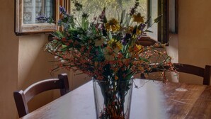 Una mesa de comedor con un jarrón de flores, una silla de madera y una ventana que ofrece vistas abiertas a una colina.