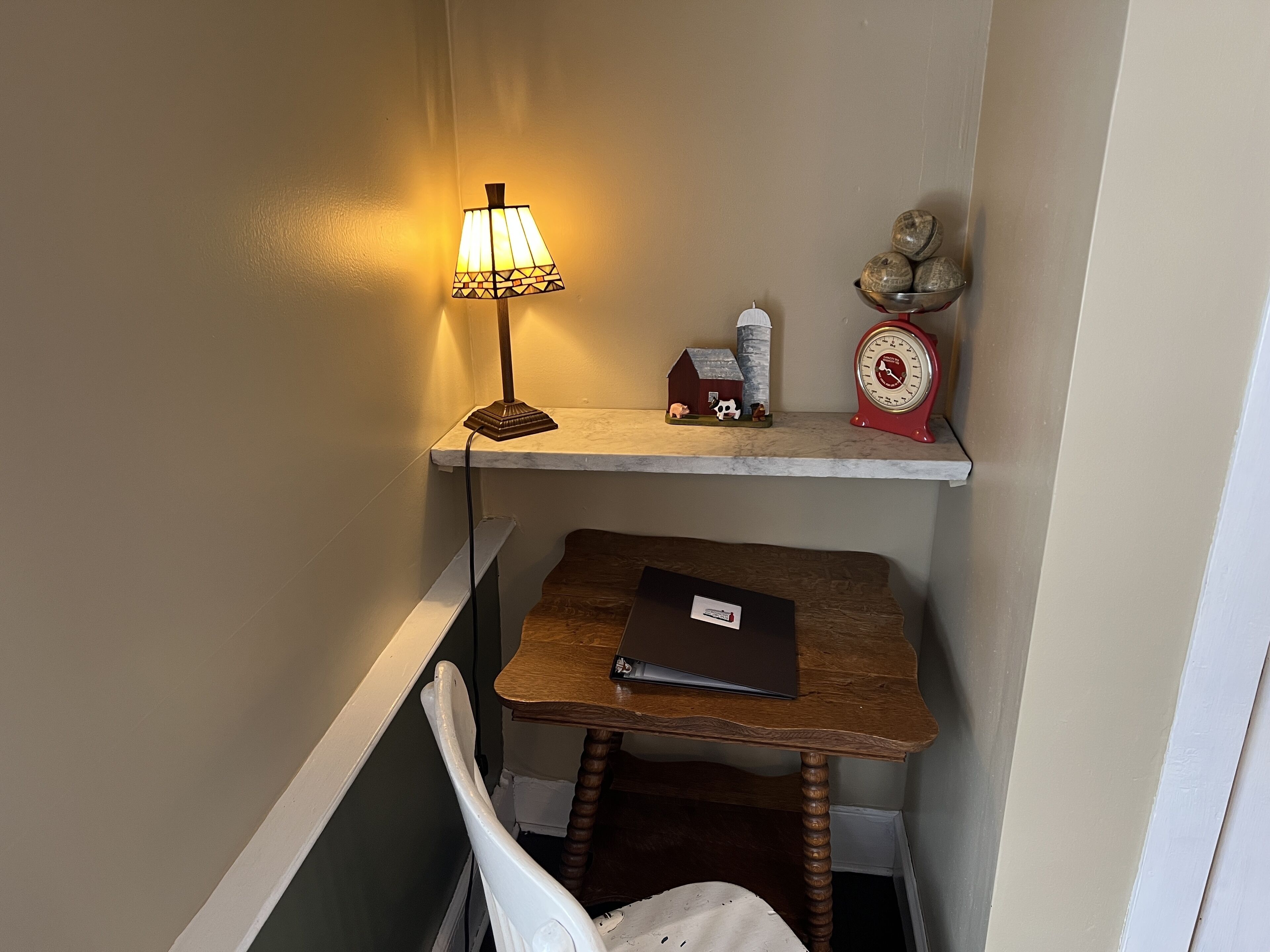 Un petit espace de travail comprenant un bureau, une chaise, une lampe, une étagère avec des objets décoratifs et une horloge.