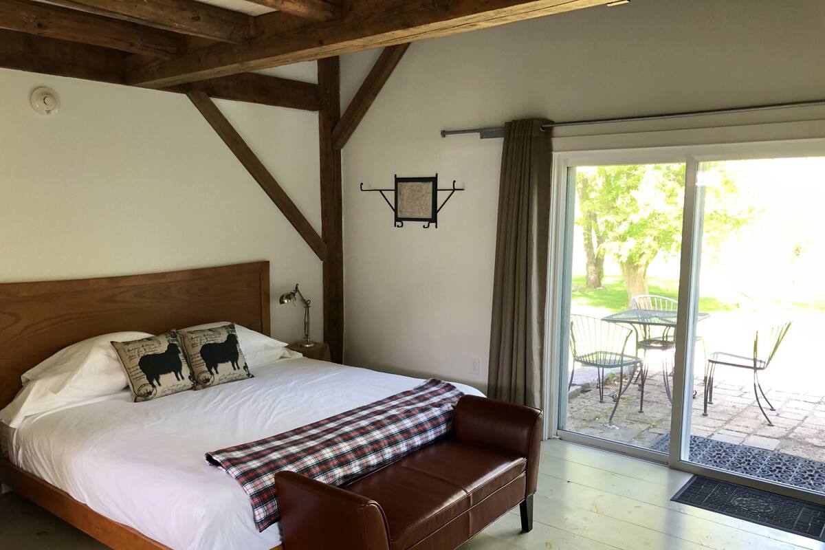 A bedroom with a wooden headboard, a bed with white linens, a brown leather bench, and a view of an outdoor patio with a table and chairs.