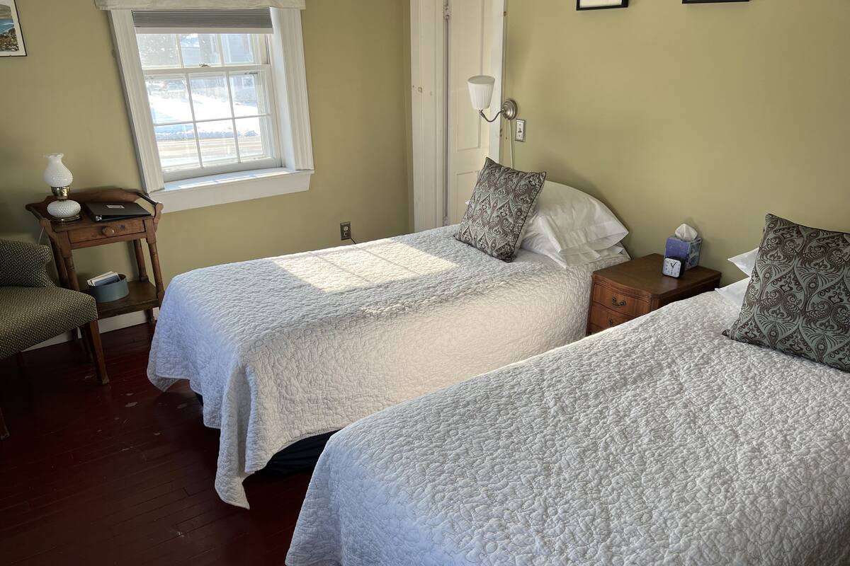A room with two beds, a nightstand with a lamp, a chair, and a window with blinds.