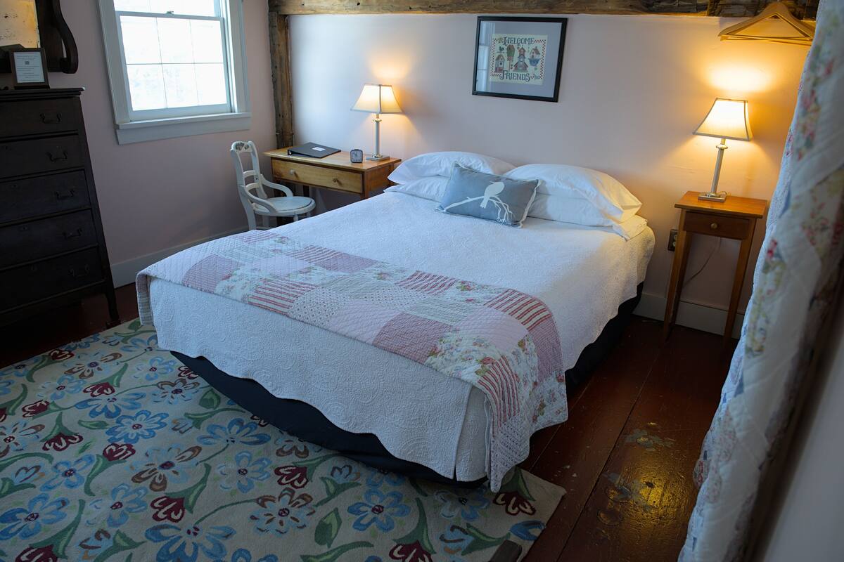 A neatly made bed with a floral bedspread, two bedside lamps, a wooden nightstand, a chair, and a dresser.
