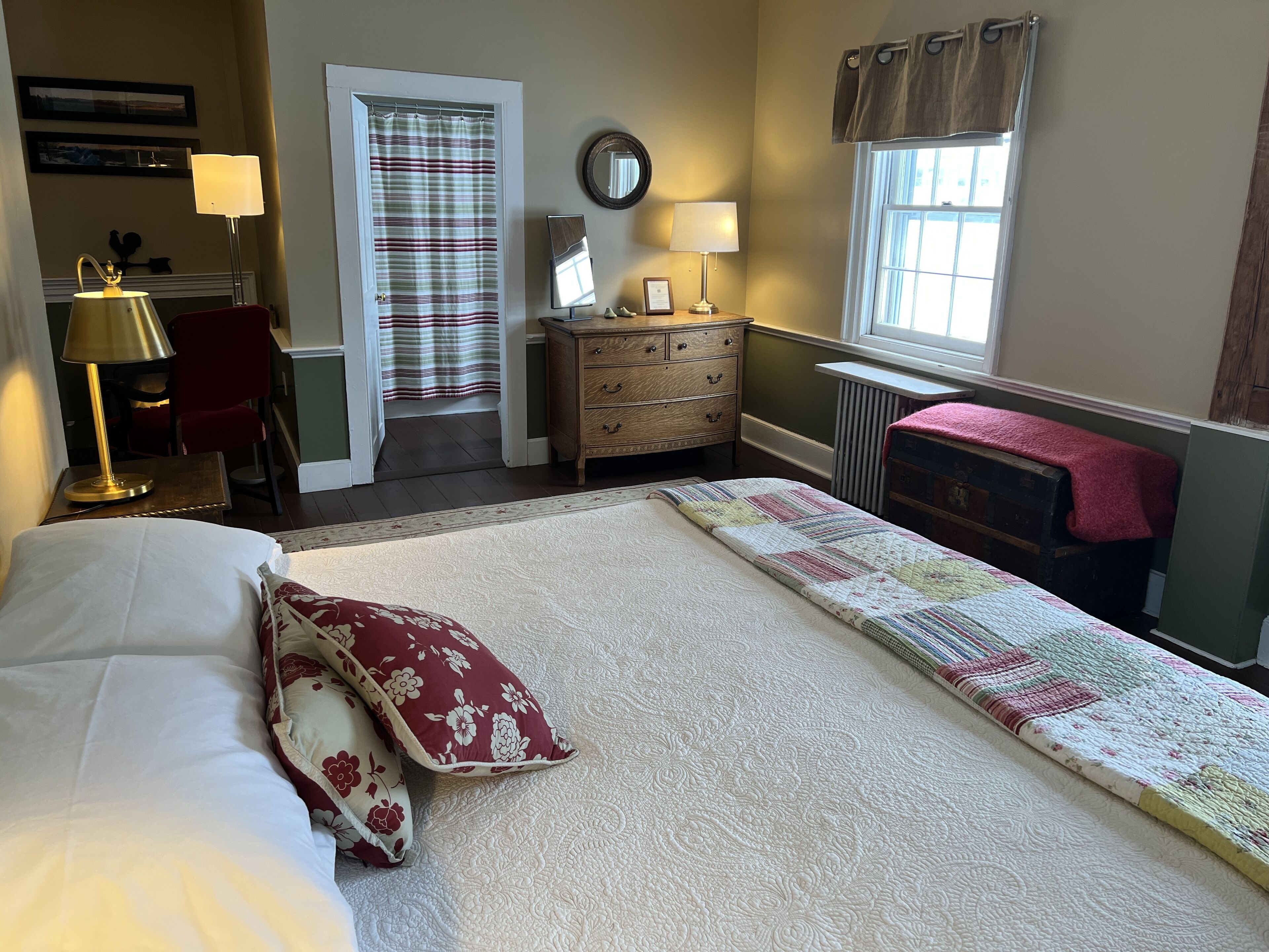 A hotel room with a bed, a desk with a lamp, a dresser, a mirror, and a window with curtains.
