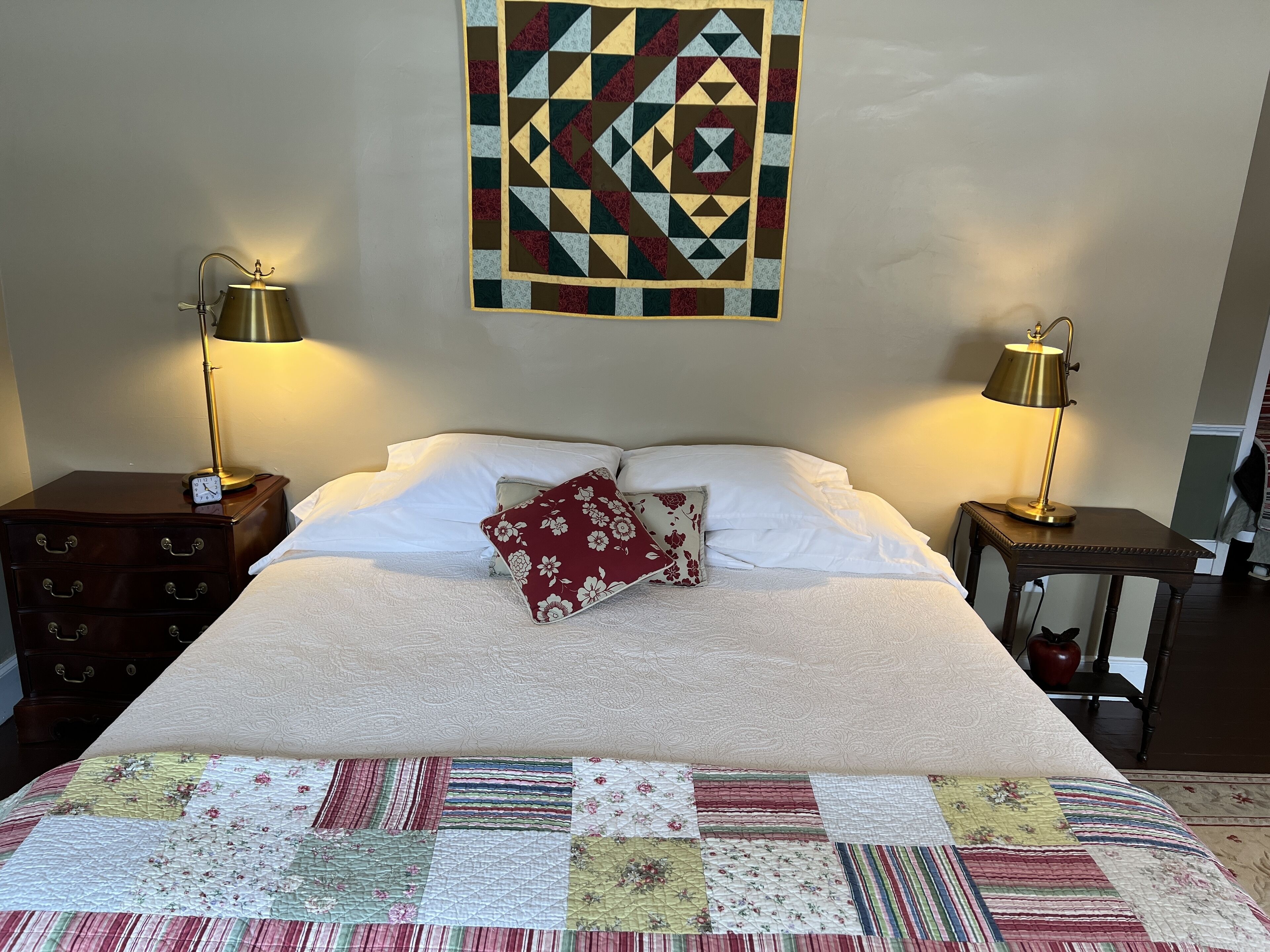 Un lit bien fait, agrémenté d’un coussin à motifs floraux, une courtepointe aux différents motifs, deux lampes de chevet, une table de chevet en bois et une décoration murale.