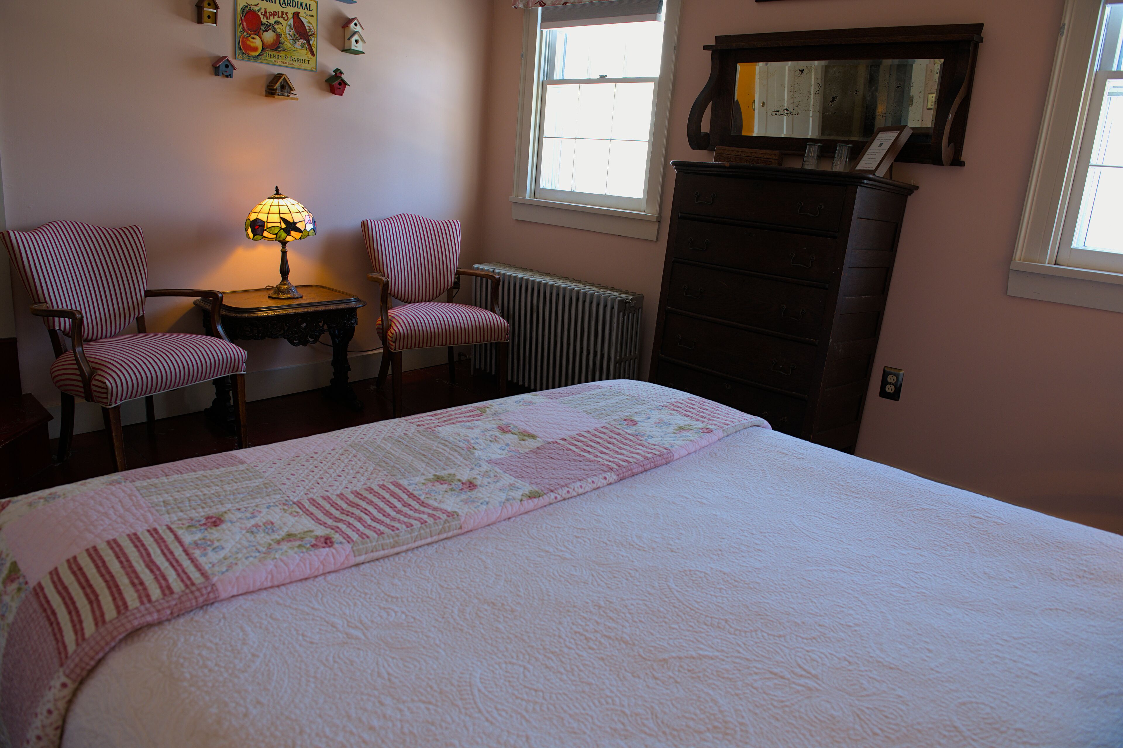 Un dormitorio con una cama grande, dos sillones a rayas, una mesa con una lámpara, un aparador y un radiador.