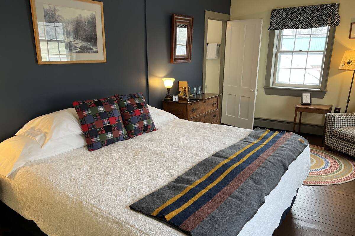 A neatly made bed with a white duvet and plaid pillows, a wooden dresser with a lamp, a framed picture on the wall, and a window with a patterned valance.