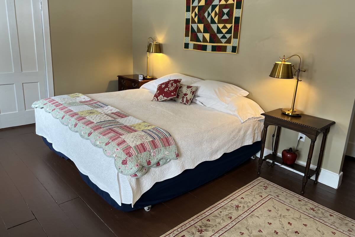 A neatly made bed with a quilt, two bedside lamps, a decorative wall hanging, and a small table with an apple.