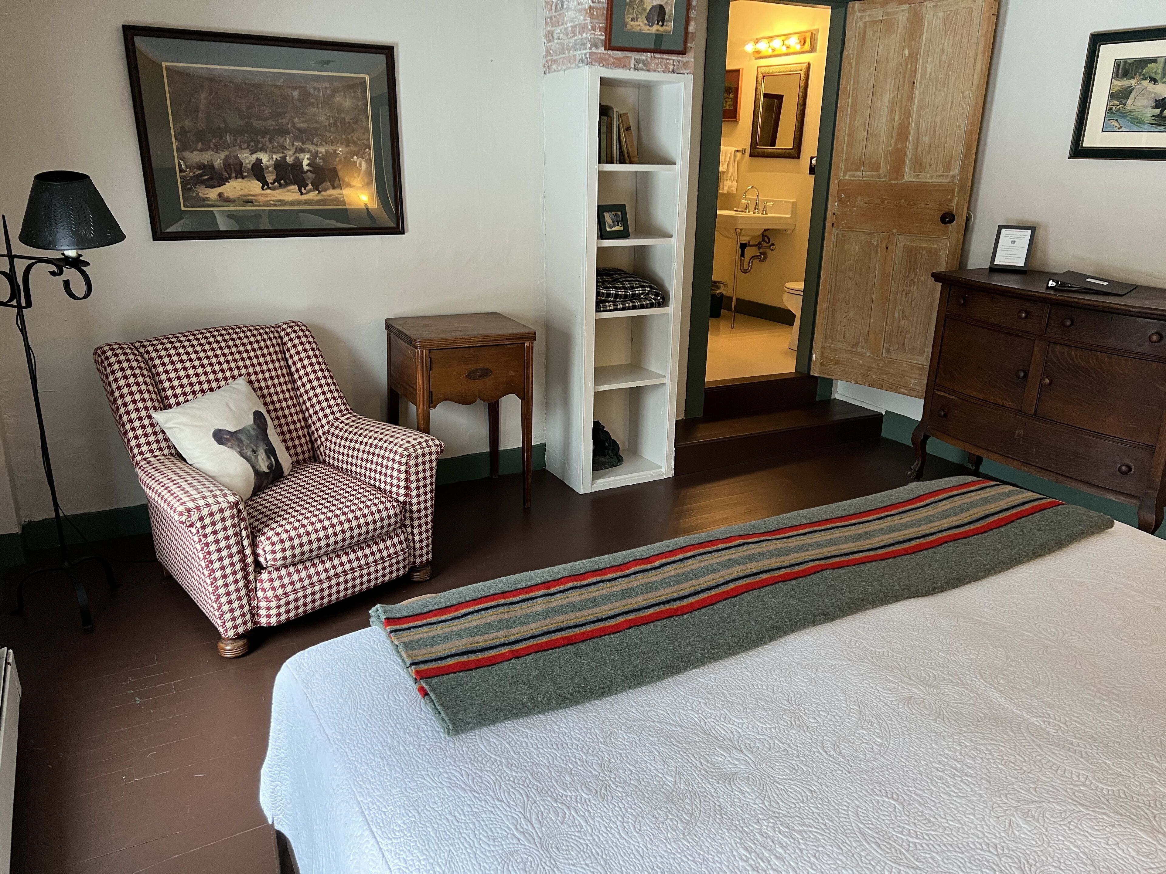 Ein Schlafzimmer mit einem Bett, einem hundszahn-gemusterten Sessel, einem kleinen Holfnachtkästchen, einem weißen Regal und einer Holzkommode.