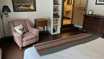 Ein Schlafzimmer mit einem Bett, einem hundszahn-gemusterten Sessel, einem kleinen Holfnachtkästchen, einem weißen Regal und einer Holzkommode.