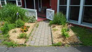En havevej der fører til en veranda med en rød bygning, omgivet af forskellige planter og blomster.