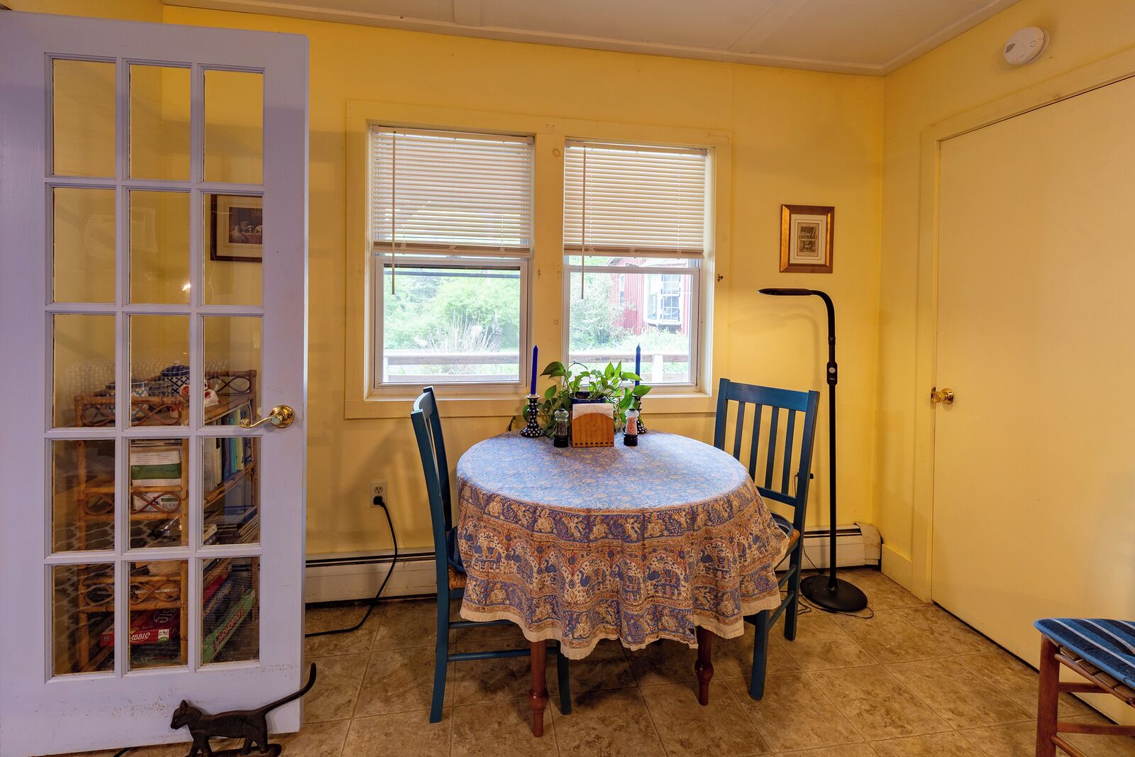 A room with a round table covered with a patterned tablecloth, two chairs, a potted plant, and a window with blinds.
