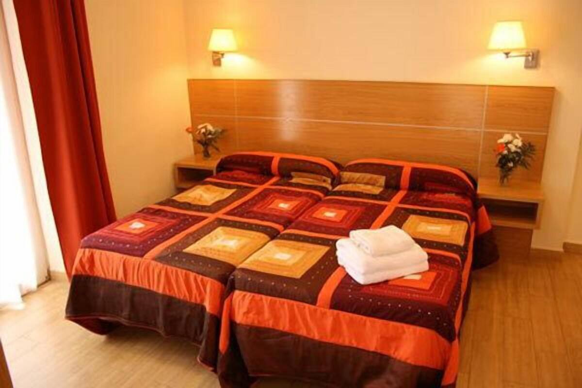 A double bed with a patterned bedspread, two bedside lamps, a nightstand with a vase, and a window with curtains.