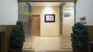 Entrada de hotel con piso de madera, puertas de vidrio y dos plantas en macetas.