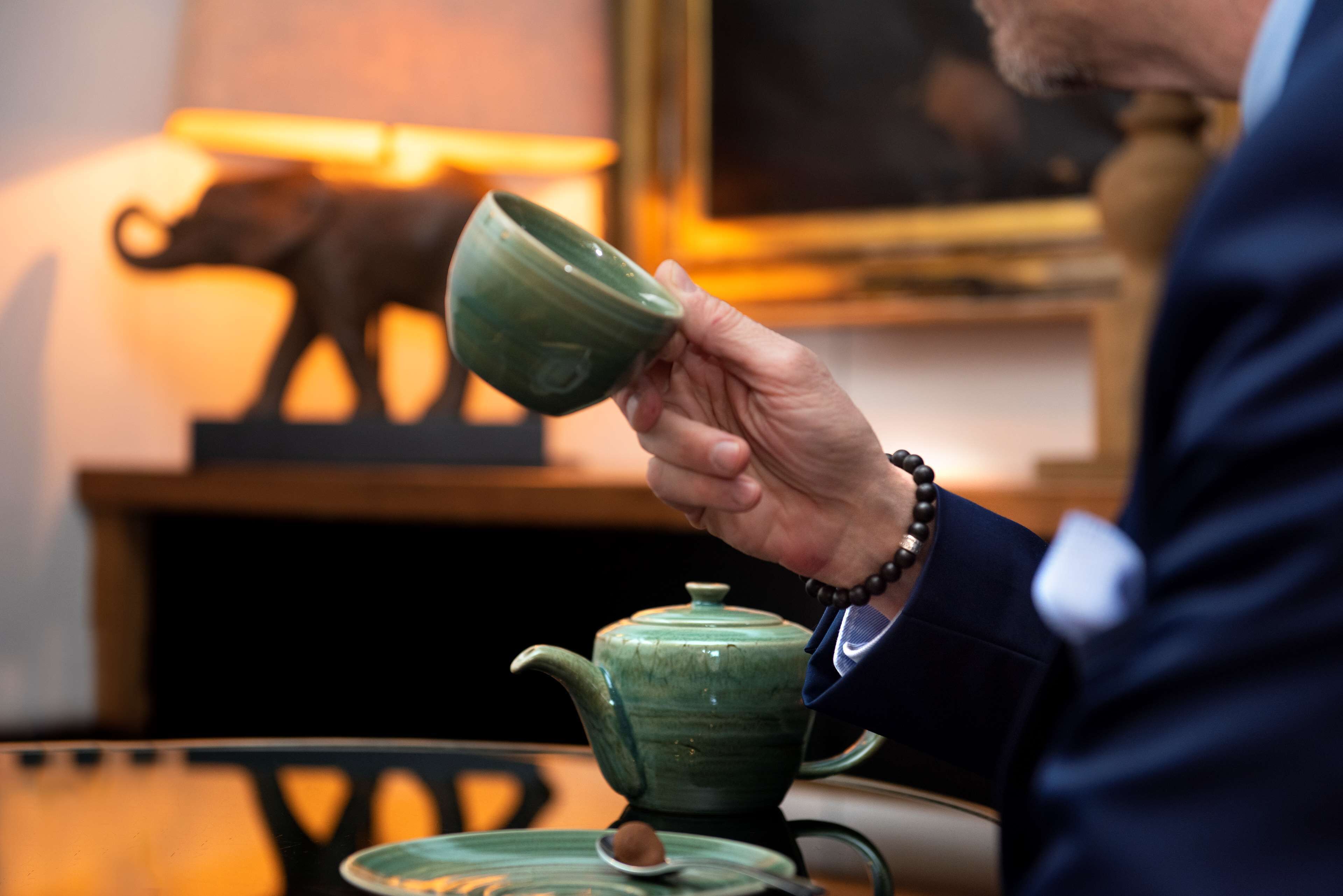 A person holding a green teapot, a spoon with a tea bag, and a teacup on a table.