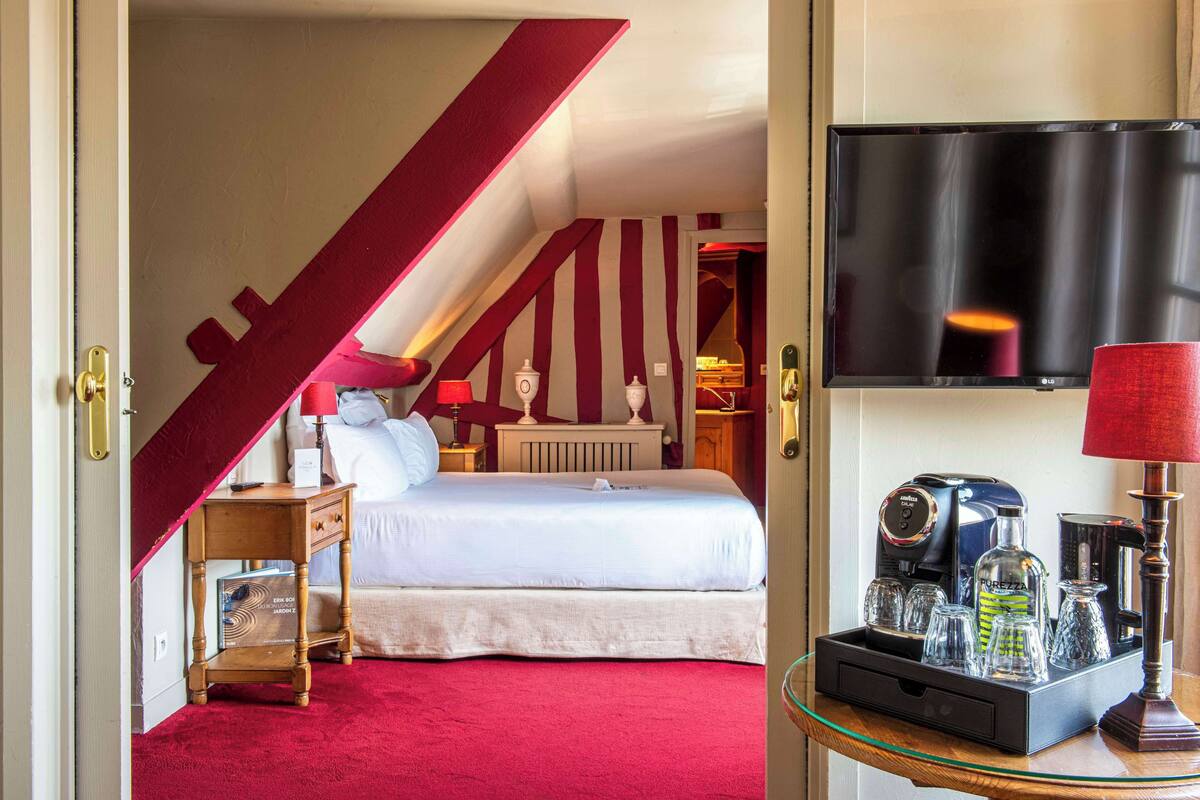 Una habitación de hotel con cama, cafetera, televisor y una mesa de vidrio con lámpara.