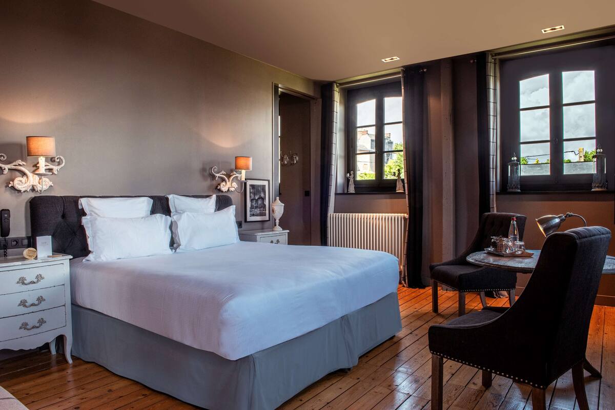 Un dormitorio con una cama grande, mesitas de noche, una silla, una mesa y una ventana con cortinas.