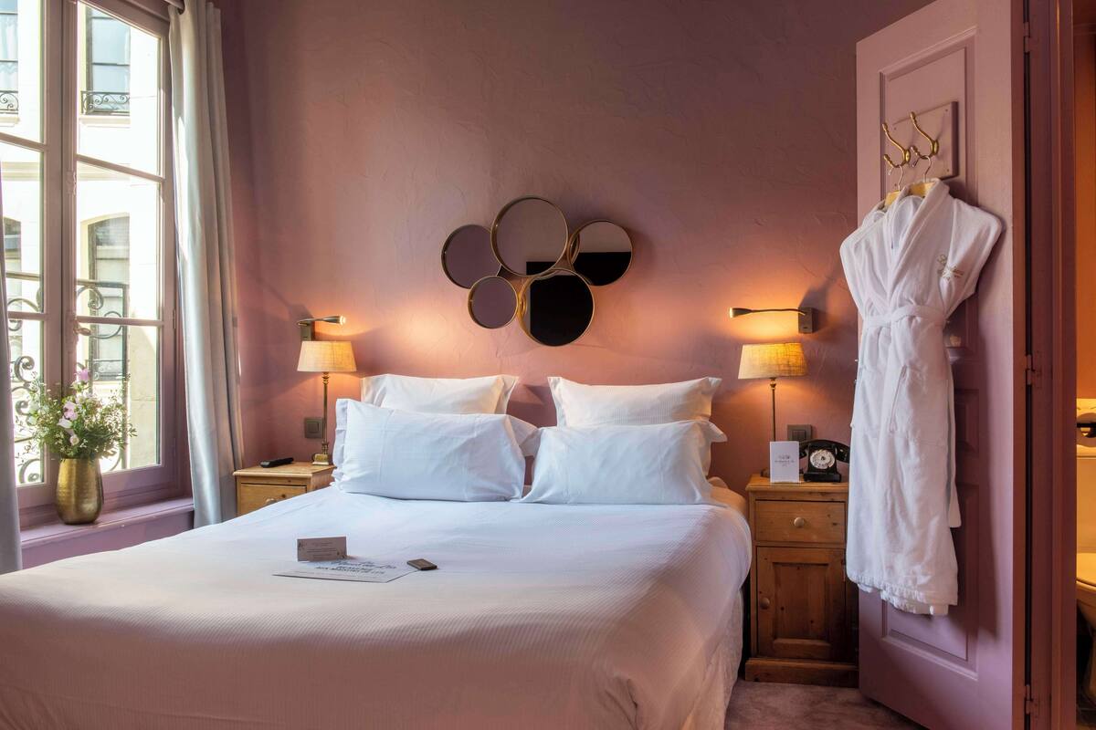 Habitación de hotel con una cama grande, lámparas de noche, un espejo en la pared, una bata colgada en la puerta y una ventana con cortinas.