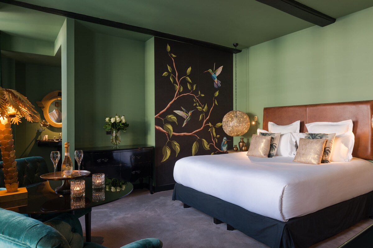 Ein Schlafzimmer mit einem großen Bett, einem Nachttisch mit Lampe, einem Sessel und einer Wand mit einem Wandbild zum Thema Natur.
