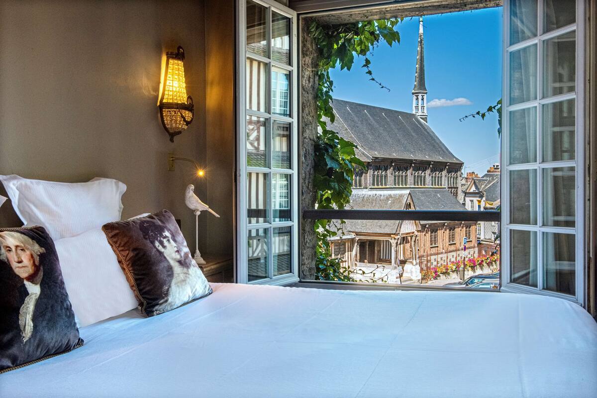 Un dormitorio con una cama grande, dos almohadas, una lámpara de pared y vista a un edificio con una torre.