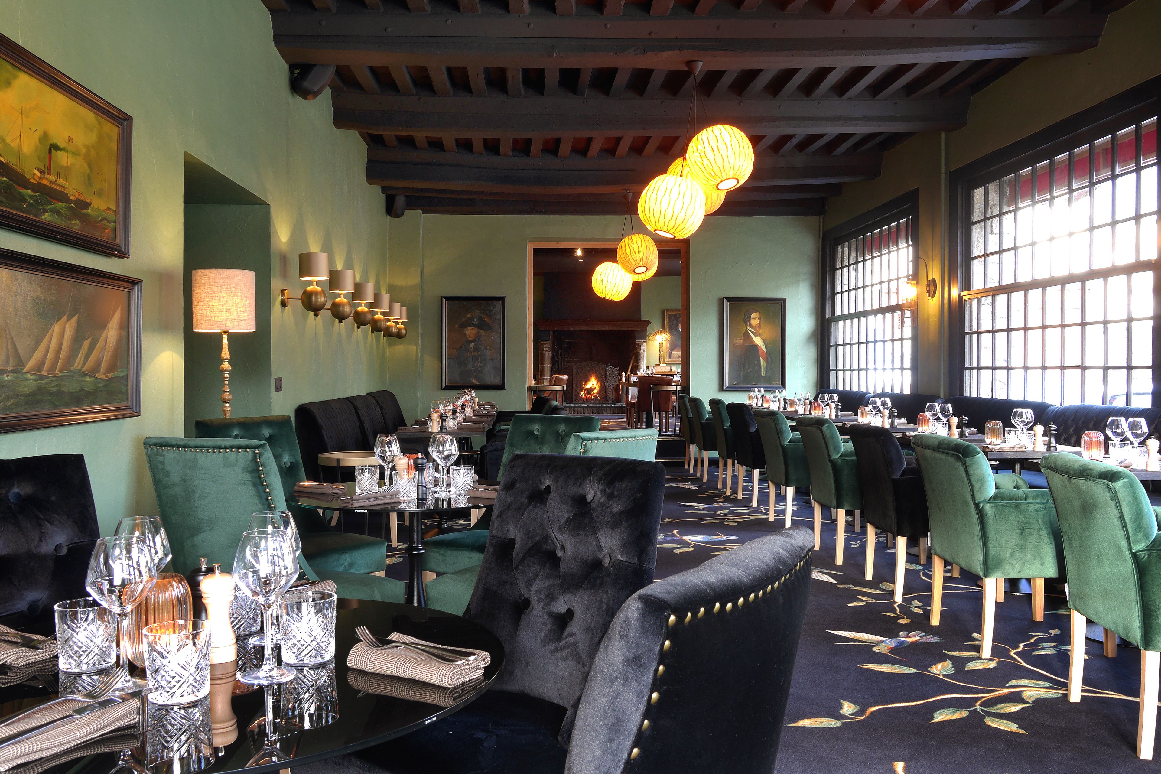 Un restaurante con sillas tapizadas de verde, una silla de terciopelo negro, y una mesa preparada con copas de vino y una vela.