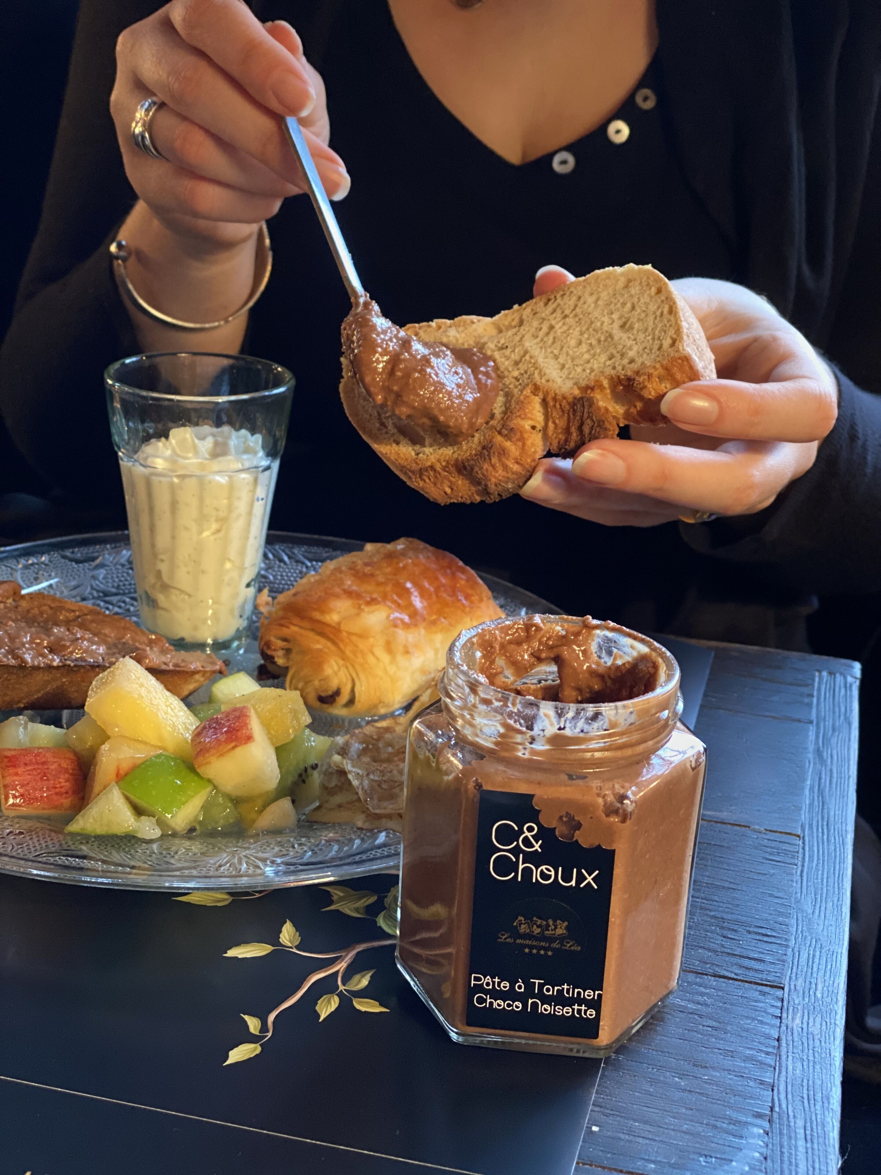 A person holding a piece of bread with a spread, a glass of milk, a jar of chocolate spread, and a plate with fruit and pastries.