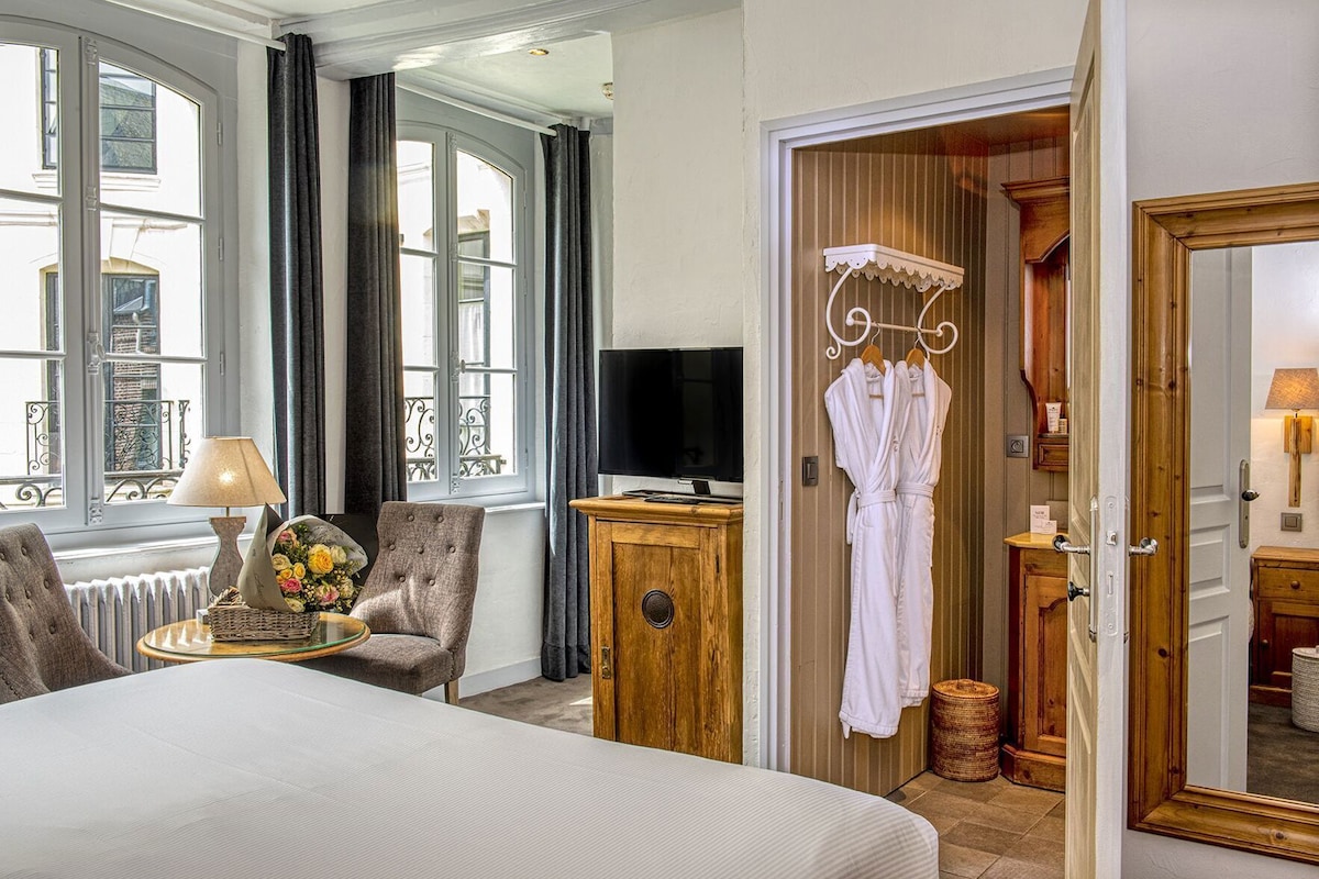 Ein Hotelzimmer mit einem Bett, einem Fernseher, einer Holzkabine, einem Sessel, einem Fenster mit Vorhängen und einer Tür mit Spiegel.