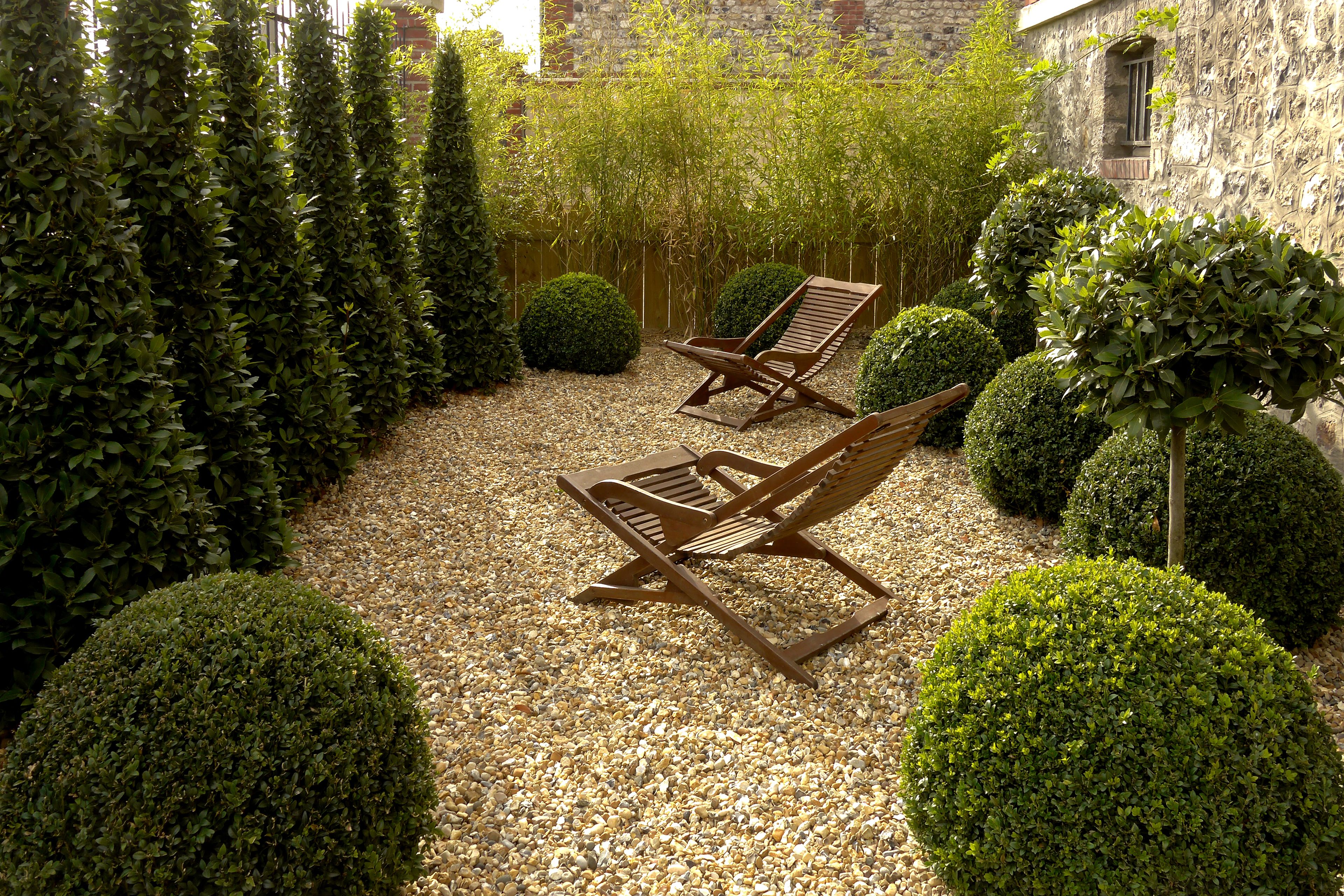 A garden with two wooden lounge chairs, neatly trimmed bushes, and a gravel path.