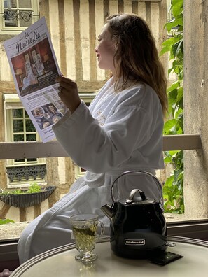 Eine Person liest eine Zeitung, gekleidet in einen weißen Bademantel, und sitzt an einem Tisch mit einem Wasserkocher und einer Teetasse.