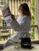 Una persona leyendo un periódico, vestida con una bata blanca, sentada a una mesa con una tetera y una taza de té.