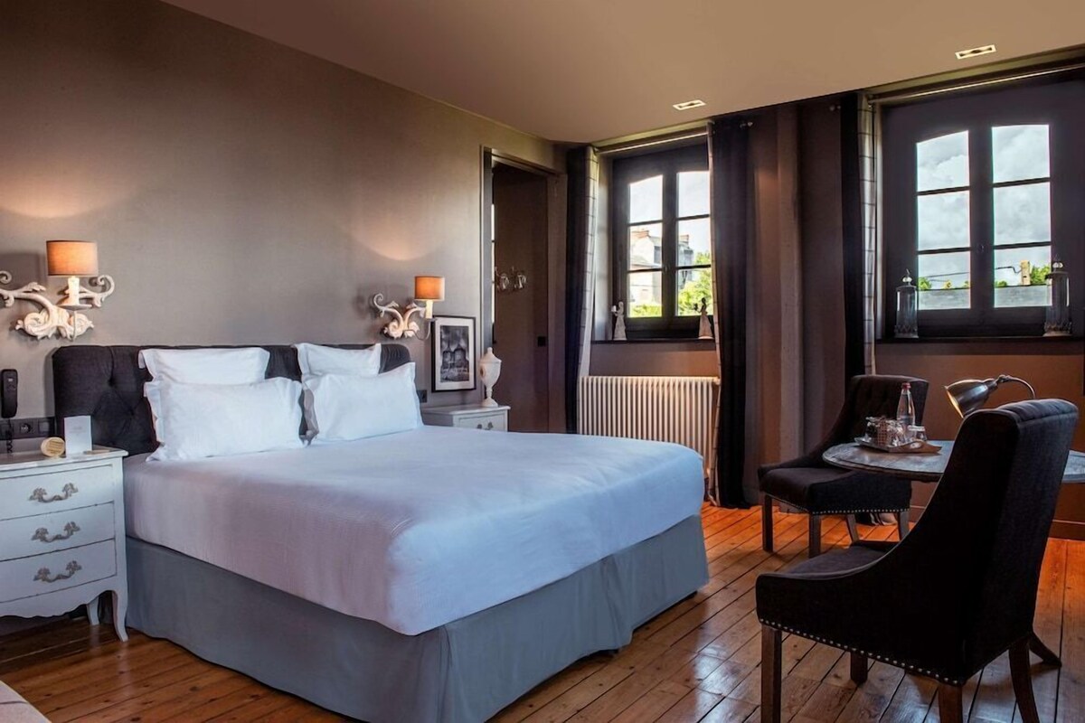 Ein Schlafzimmer mit einem großen Bett, Nachttischen, einem Sessel, einem kleinen Tisch und einem Fenster mit Vorhängen.