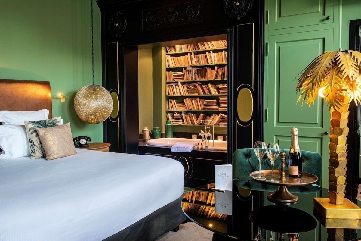 Ein Schlafzimmer mit einem Bett, einer Bücherwand, einem Schminktisch, einem grünen Sessel und einem kleinen Tisch mit Gläsern und einer Flasche.